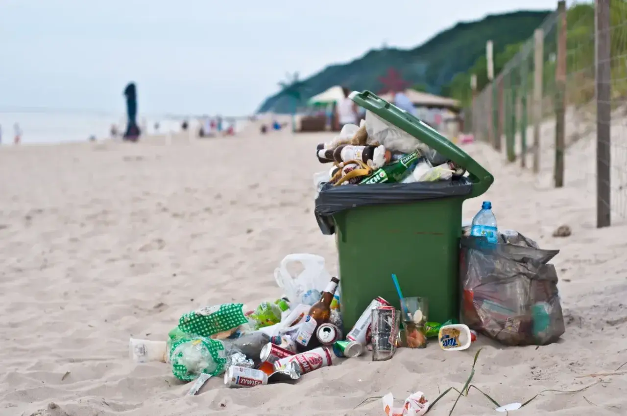 Przepełniony kosz i śmieci na plaży to przyczyny zanieczyszczenia Bałtyku. Butelki, puszki i plastik zalegają na piasku.