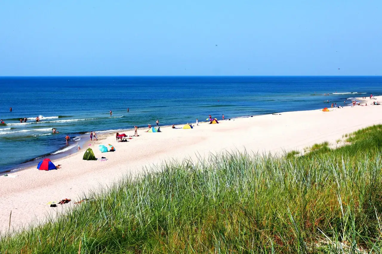 Słoneczny dzień nad Bałtykiem, ludzie kąpią się w wodzie, ciesząc się przyjemną temperaturą. Na plaży rozstawione namioty i parawany.