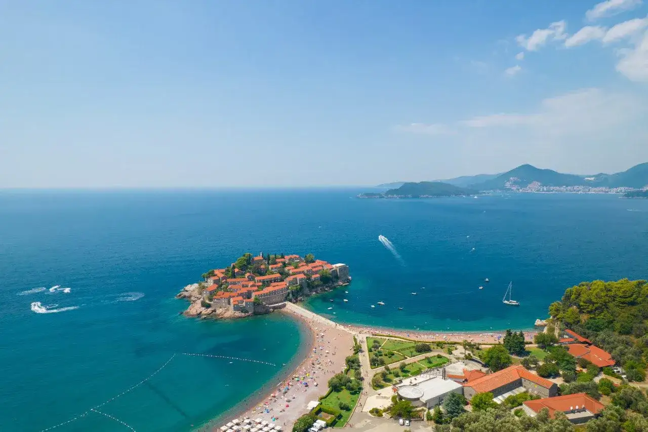 Czarnogóra jakie morze? Piękna wyspa Sveti Stefan z czerwonymi dachami, otoczona turkusowym Adriatykiem, z plażą pełną ludzi i górami w tle.