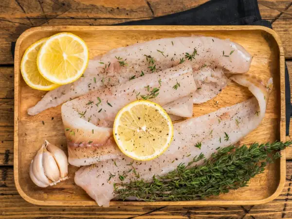 Filety z mintaja na drewnianej desce, przyprawione ziołami i pieprzem, z cytryną i czosnkiem. Czy mintaj ma ości? Te filety wyglądają na pozbawione ości.