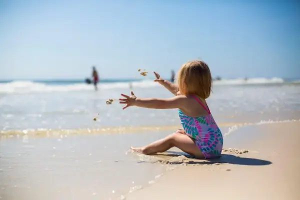 Dziewczynka w kolorowym stroju kąpielowym rzuca piasek na plaży. Idealny obrazek do poradnika: co zabrać na wakacje nad morze z dzieckiem.