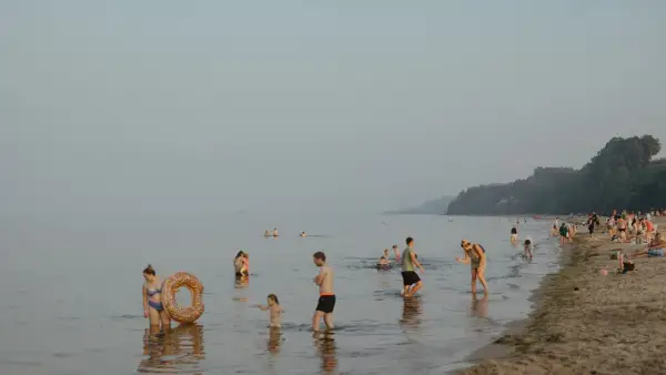 Ludzie kąpią się i spacerują po plaży, podczas gdy nad morzem unosi się delikatna mgła. W oddali widać statek.