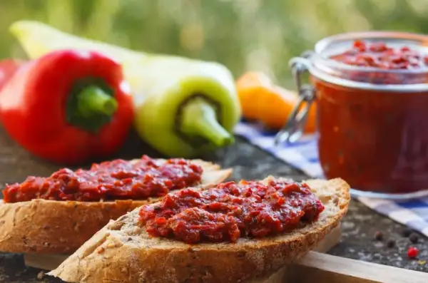 Chleb z pyszną pastą z pieczonej papryki, obok słoiczek i świeże papryki.