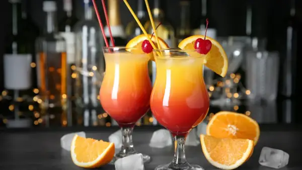 Dwa drinki w domu, ozdobione pomarańczą i wiśnią, z kostkami lodu i owocami w tle.